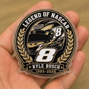 Op de afbeelding: Een cirkelvormig gedenkitem met de tekst "LEGEND OF NASCAR" en "KYLE BUSCH 1985-2026". Het ontwerp toont een racehelm met het nummer 8 en het M&M's-logo, in zwart, goud en wit.