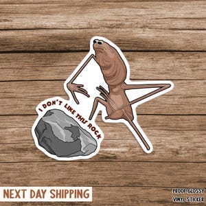 May include: A glossy vinyl sticker showing a cartoon alien with long limbs and a slender body, looking displeased at a grey rock. The text "I DON'T LIKE THIS ROCK" is included. The sticker is on a wooden background.