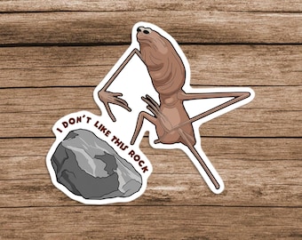 May include: A glossy vinyl sticker featuring a cartoon alien with long limbs and a slender body, looking displeased at a gray rock. The text "I DON'T LIKE THIS ROCK" is included. The sticker is on a wooden background.
