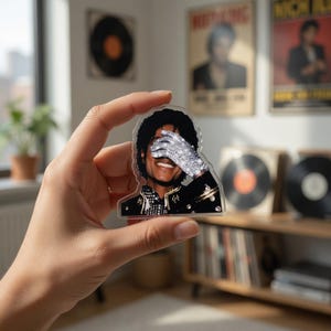 Könnte beinhalten: Ein klarer Acrylaufkleber mit einer lächelnden Person, die sich die Augen mit einem glitzernden Handschuh bedeckt. Die Person trägt eine schwarze Jacke mit goldenen Akzenten. Der Aufkleber wird vor einem verschwommenen Hintergrund mit Vintage-Albumcovern gehalten.