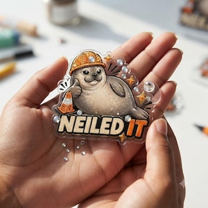 May include: A clear acrylic badge featuring a cartoon seal wearing an orange construction hard hat and giving a thumbs up next to a traffic cone. The text NEILED IT is printed in bold letters at the bottom, accented with small clear rhinestones.