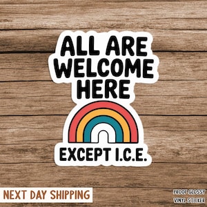 May include: A white sticker with the text "ALL ARE WELCOME HERE EXCEPT I.C.E." above a rainbow design. The sticker has a glossy finish and is on a wooden background.