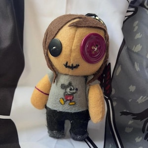 May include: A handmade doll with button eyes and stitched mouth. The doll has brown hair, a gray t-shirt with a Mickey Mouse graphic, and black pants. A key ring is attached to the top of the doll's head. The doll is made of felt.