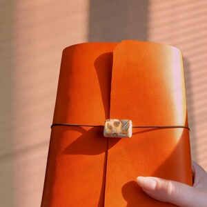 May include: An orange leather journal with a decorative clasp and a dark brown cord closure. The journal is held by a person's hand. The background is a soft peach color.