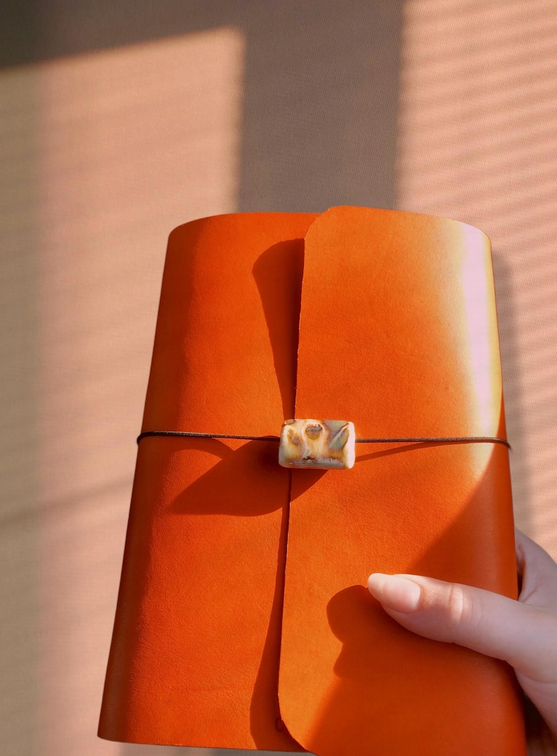 May include: An orange leather journal with a decorative clasp and a dark brown cord closure. The journal is held by a person's hand. The background is a soft peach color.