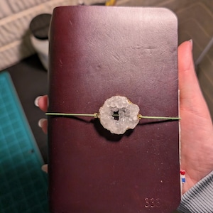 May include: A dark burgundy leather journal with a geode accent. The geode is white with gold-colored trim and is held in place by a green elastic band. The journal has the number "S36" embossed on the front.