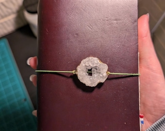 May include: A dark burgundy leather journal with a geode accent. The geode is white with gold-colored trim and is held in place by a green elastic band. The journal has the number "S36" embossed on the front.