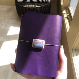 May include: A purple leather journal with a decorative clasp and a green elastic closure. The clasp is a rectangular bead with pastel colors. The journal is held in a person's hand.
