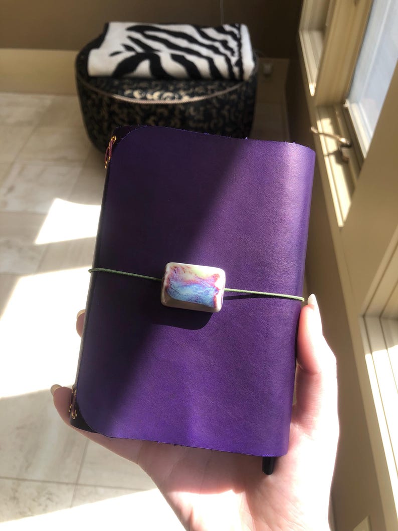 May include: A purple leather journal with a decorative clasp and a green elastic closure. The clasp is a rectangular bead with pastel colors. The journal is held in a person's hand.