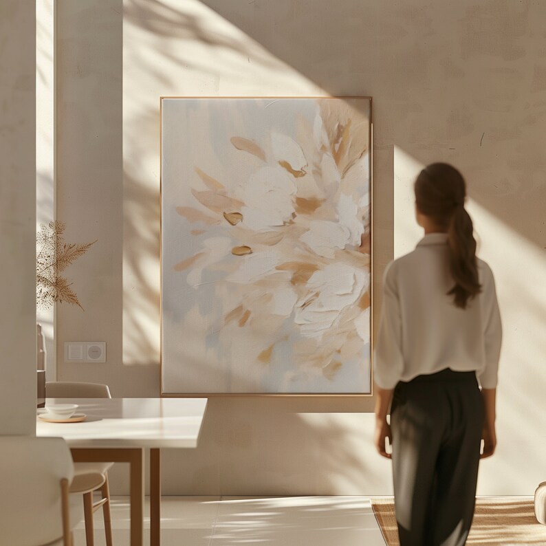 May include: A large abstract painting with a light blue, white, and beige color scheme. The painting is framed in a thin, gold frame and hangs on a light beige wall. A person is standing in front of the painting, looking at it.