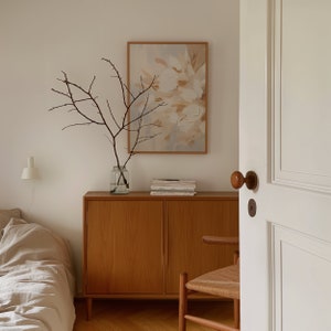 May include: A wooden cabinet with two doors and a light wood finish. The cabinet is decorated with a vase of dried branches and a framed abstract painting with beige and white colors.