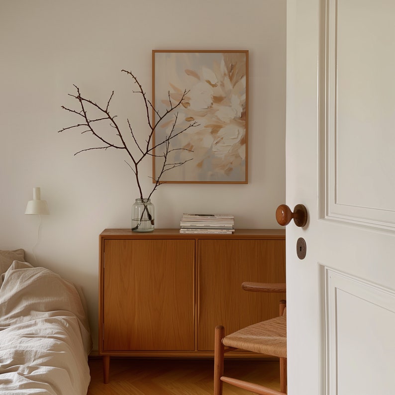 May include: A wooden cabinet with two doors and a light wood finish. The cabinet is decorated with a vase of dried branches and a framed abstract painting with beige and white colors.