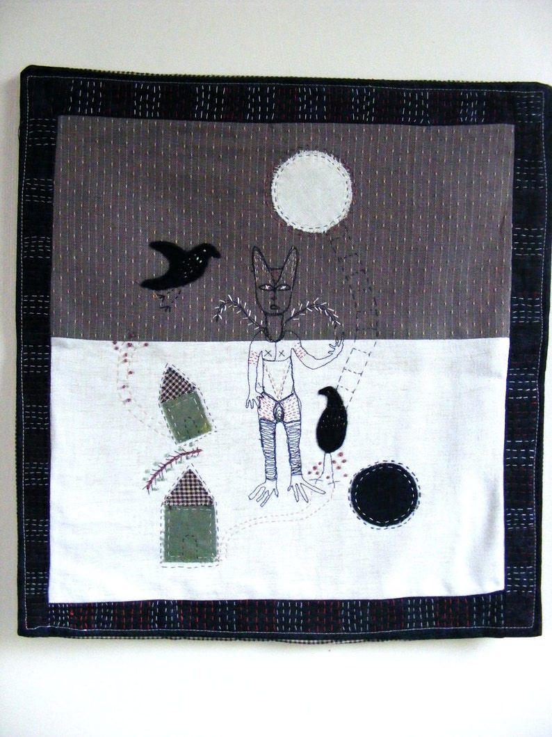 May include: A black and white embroidered wall hanging with a stylized figure, two birds, and geometric shapes. The background is a dark gray fabric with a white moon.