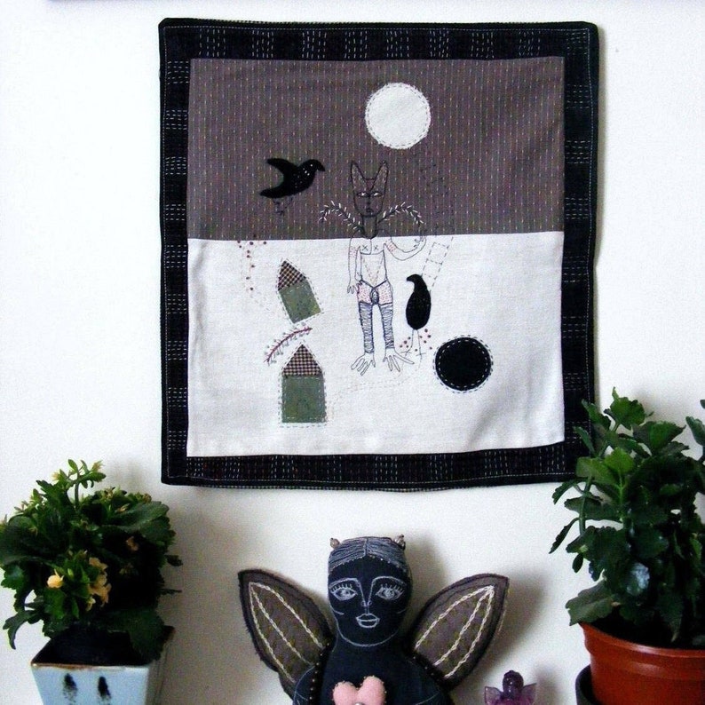 May include: A black and white embroidered wall hanging with a brown background. The embroidery depicts a figure with a wolf head, a bird, and two small houses. The hanging is framed with a black border.