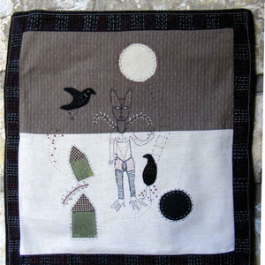 May include: A brown and white fabric wall hanging with a black bird, a human figure, and two small houses. The background is a brown fabric with a white circle in the upper center. The bottom half of the wall hanging is white fabric.