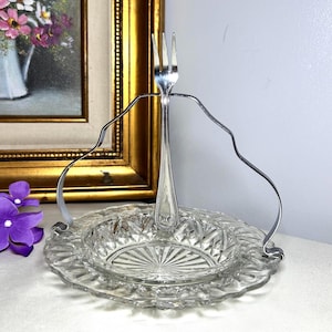 May include: A clear glass serving dish with a decorative silver handle and a fork. The dish has a scalloped edge and a diamond pattern. The fork is upright in the center of the dish. A gold framed painting is in the background.