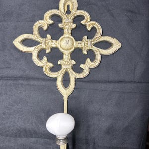 May include: Cream-colored wall hook with an ornate, fleur-de-lis design. The hook features a white, oval-shaped knob at the bottom. The metal has a distressed finish, adding a vintage aesthetic.
