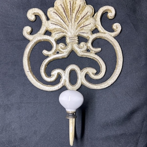 May include: Cream-colored wall hook with a decorative, ornate design. The hook features a stylized shell-like top and a white, round knob. The hook is made of metal and has a vintage, antique appearance. The hook is designed for hanging items.