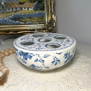 May include: A white ceramic flower frog bowl featuring a blue floral pattern. The top surface has five large circular openings surrounded by smaller holes for stems. The bowl has a rounded shape and is decorated with blue vines and blossoms.
