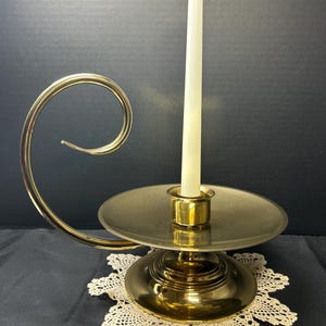 May include: A gold-colored candle holder with a curved handle and a circular base. A white candle is inserted in the center. The candle holder is placed on a white lace doily.