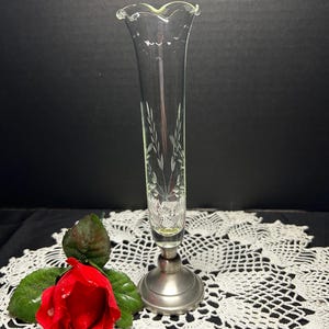 May include: A clear glass bud vase with etched floral designs, set on a silver-colored metal base. The vase has a ruffled top edge. A red rose with green leaves rests nearby.