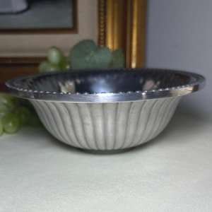 Op de afbeelding: Een zilverkleurige kom met een gegroefde buitenkant en een kralenrand. De kom is rond en staat op een lichtgekleurd oppervlak. Groene druiven en een schilderij staan op de achtergrond.