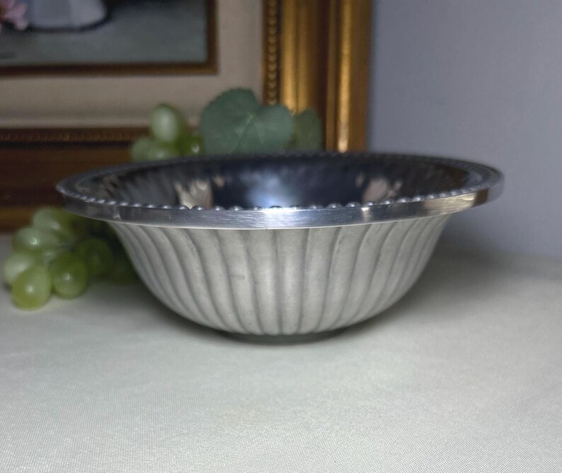 Op de afbeelding: Een zilverkleurige kom met een gegroefde buitenkant en een kralenrand. De kom is rond en staat op een lichtgekleurd oppervlak. Groene druiven en een schilderij staan op de achtergrond.