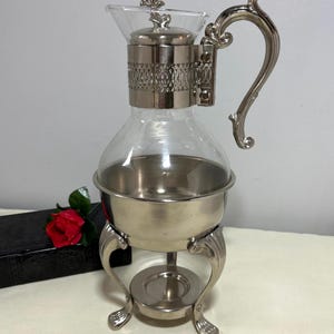 May include: A clear glass carafe with a silver-toned metal base and a matching silver-toned metal collar featuring a decorative lattice pattern. The carafe has a curved handle, a metal lid with a small finial, and stands on three ornate metal legs.