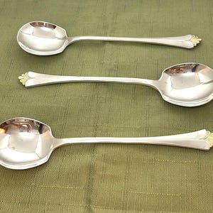 May include: Three silver-toned spoons with polished, reflective bowls and handles. Each handle features a distinct gold-toned, fan-shaped decorative accent at the tip. The spoons are arranged in a parallel formation, showcasing a smooth, metallic finish throughout the stems and bowls.