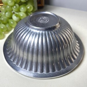 Op de afbeelding: Een vintage zilverkleurige metalen kom met een gegroefd ontwerp. De kom heeft een afgeronde vorm met een brede, platte basis en een decoratieve rand. De bodem van de kom heeft een cirkelvormig embleem. De kom wordt waarschijnlijk gebruikt om te serveren of tentoon te stellen.