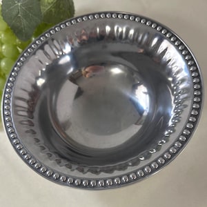 Op de afbeelding: Een glanzende, zilverkleurige kom met een kralenrand. De kom heeft een gehamerde textuur en een glad, reflecterend interieur. De rand is versierd met kleine, gelijkmatig verdeelde kralen. De kom wordt waarschijnlijk gebruikt om te serveren of tentoon te stellen.