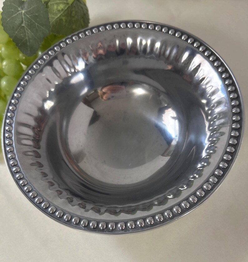 Op de afbeelding: Een glanzende, zilverkleurige kom met een kralenrand. De kom heeft een gehamerde textuur en een glad, reflecterend interieur. De rand is versierd met kleine, gelijkmatig verdeelde kralen. De kom wordt waarschijnlijk gebruikt om te serveren of tentoon te stellen.