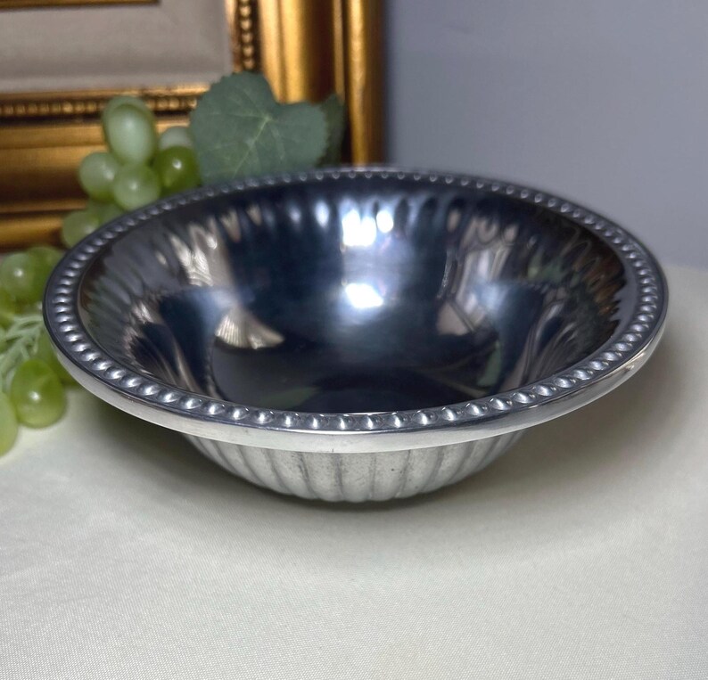 Op de afbeelding: Een kleine, ronde, zilverkleurige kom met een kralenrand en een gegroefde buitenkant. De binnenkant van de kom is glanzend en reflecterend. De achtergrond bevat een goudkleurige lijst en groene druiven.