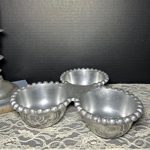May include: A three-sectioned, silver-colored serving dish with a beaded edge. The dish is made of metal and has a vintage aesthetic. The three bowls are connected and sit on a lace tablecloth. A candle holder and a red candle are in the background.