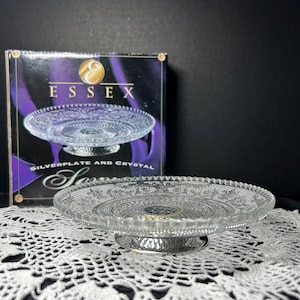 May include: Clear glass cake stand with a silver-plated base. The stand has a decorative, etched design on the plate and base. The box behind the stand reads "Essex Silverplate and Crystal".