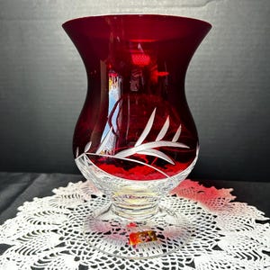 May include: A ruby red crystal vase with a flared top and a clear, etched base. The vase features a delicate white leaf design around the middle. The vase is displayed on a white lace doily.