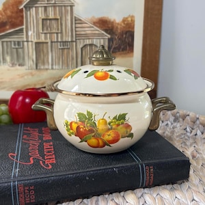 May include: A cream-colored enamel pot with a matching lid, featuring a colorful fruit motif of apples, pears, and grapes. The pot has two brass-toned metal handles and a matching brass-toned knob on the lid, with a metallic rim around the top edge.