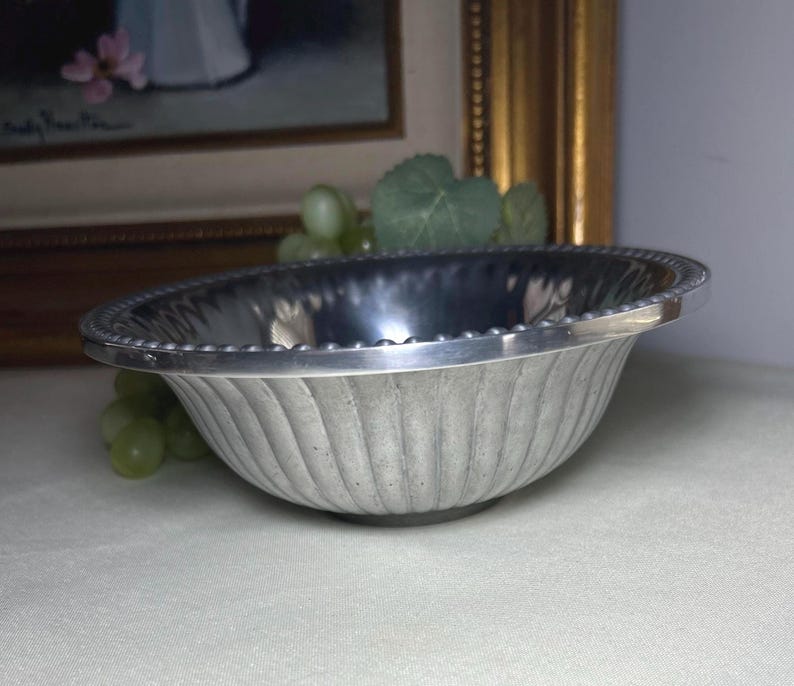 Op de afbeelding: Een zilverkleurige, ovale kom met een gegroefde buitenkant en een kralenrand. De kom staat op een wit oppervlak. De achtergrond omvat een ingelijst schilderij en groene druiven.