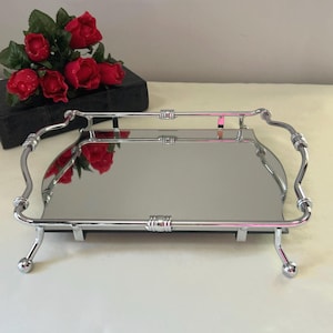 May include: A rectangular mirrored vanity tray featuring a polished chrome metal frame with curved edges, decorative cylindrical accents, and four raised legs with ball feet. The reflective surface shows a clear view of the surrounding area.
