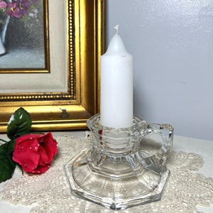 May include: A clear glass candle holder with a white candle. The candle holder has a handle and a hexagonal base. A red rose and a framed painting are in the background.