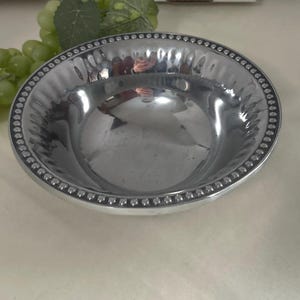 Op de afbeelding: Een glanzende, zilverkleurige kom met een kralenrand. De kom heeft een gehamerde textuur aan de binnenkant en een glad, reflecterend oppervlak. De kom is rond en lijkt van metaal te zijn. Een tros groene druiven en een blad staan op de achtergrond.