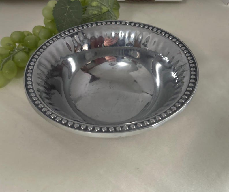 Op de afbeelding: Een glanzende, zilverkleurige kom met een kralenrand. De kom heeft een gehamerde textuur aan de binnenkant en een glad, reflecterend oppervlak. De kom is rond en lijkt van metaal te zijn. Een tros groene druiven en een blad staan op de achtergrond.