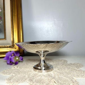 May include: A silver-colored, footed bowl with a textured floral pattern on the interior. The bowl has a wide, shallow design and a pedestal base. The background includes a gold-framed painting and purple flowers.