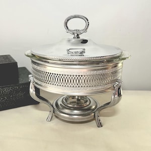 May include: A silver-plated chafing dish with a lid and a decorative handle. The dish has a glass insert and sits on a stand with curved legs. The design includes an openwork pattern around the body.