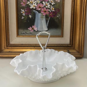 May include: A white, two-tiered serving dish with a ruffled edge. It has a silver handle in the center. The background features a framed painting of flowers in a vase.