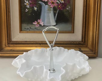 May include: A white milk glass serving dish with a ruffled edge and a central silver-toned metal handle. The dish features a circular base and a decorative, scalloped rim, providing a classic look for serving small items or displaying decorative objects.