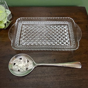 May include: A rectangular clear glass serving tray with a diamond-patterned base and integrated side handles, paired with a silver-toned metal serving spoon featuring a circular bowl with intricate floral cutouts and a smooth handle.
