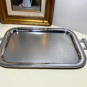 May include: A rectangular silver-toned serving tray with a polished metallic finish and a textured border. The tray features two decorative handles on opposite sides, each with a geometric pattern, and a smooth, flat central surface area.
