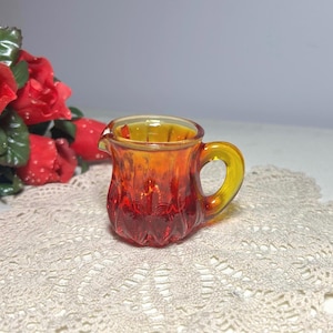 May include: A small, vintage glass creamer with a handle and spout. The creamer is a gradient of orange and yellow, with a textured, ribbed design. The handle is a bright yellow color.