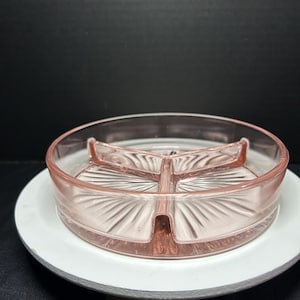 May include: A vintage, pink glass divided serving dish with a circular shape. The dish has three compartments and a decorative, fan-like pattern. It sits on a white, round base.